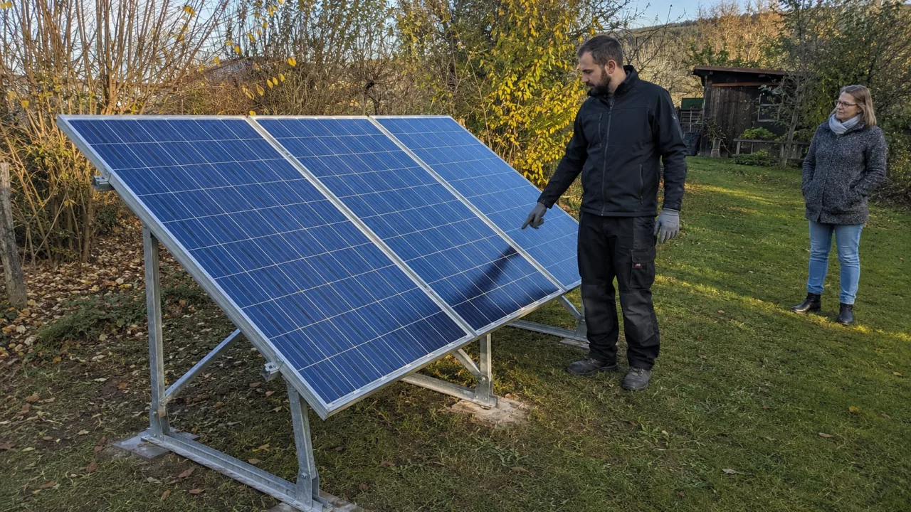 Eine Fachkraft prüft vom Boden aus drei ältere blaue Solarmodule auf einem niedrigen Metallgestell.