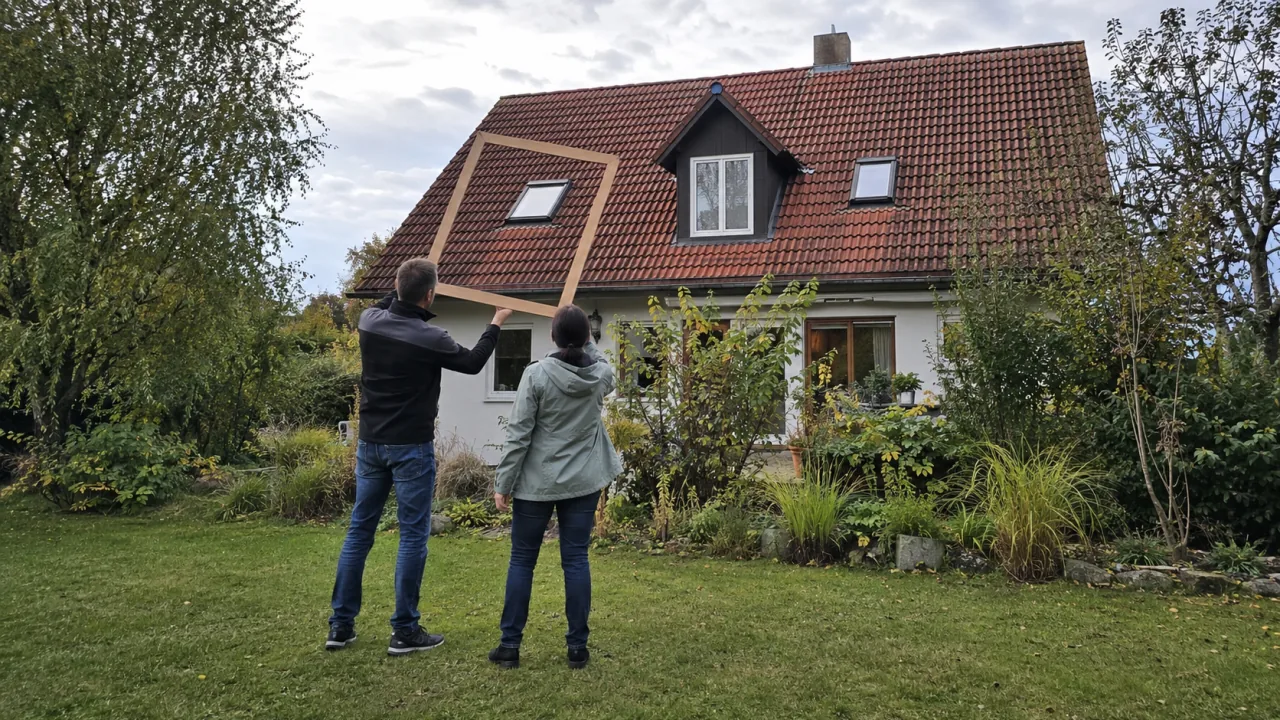Zwei Personen prüfen vom Garten aus ein Wohnhausdach mit Gaube und Dachfenstern anhand eines leeren Maßrahmens.
